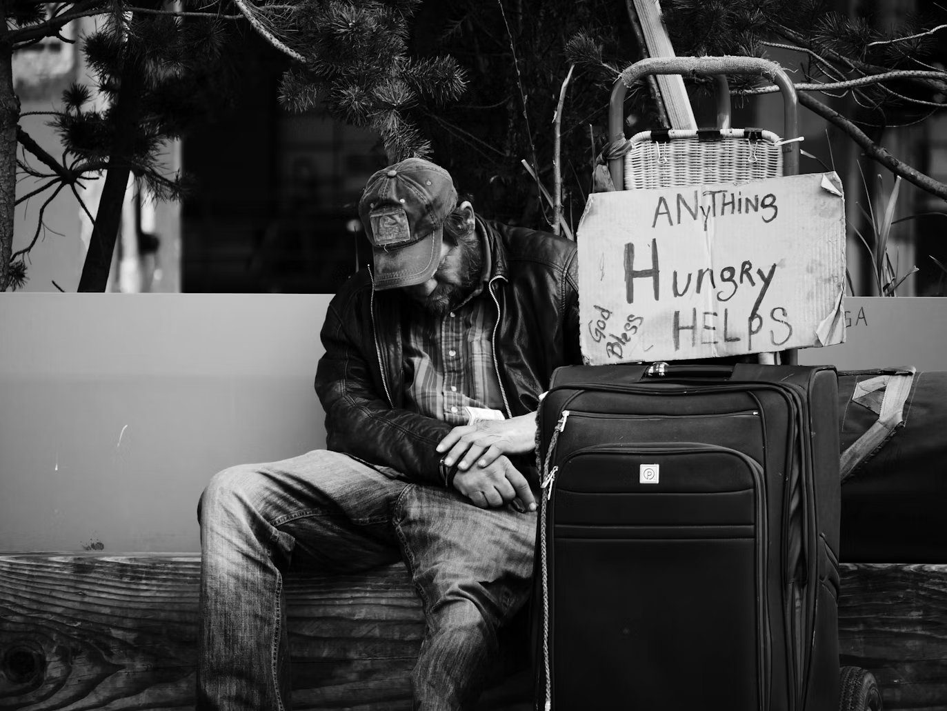 A man experiencing homelessness rests beside his belongings and a handwritten sign reading Hungry, Anything Helps, God Bless.